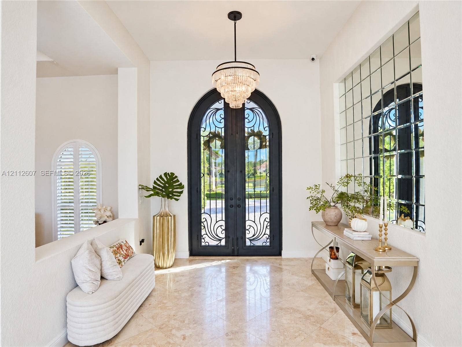 14443 S Jockey Circle S, Davie, FL 33330 Foyer featuring arched double iron entry doors, light-toned stone flooring, a multi-tiered crystal chandelier, and an expansive grid-pattern wall mirror