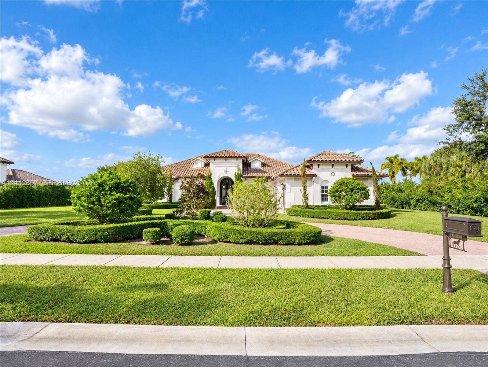 14443 S Jockey Circle S, Davie, FL 33330 Stucco exterior featuring a clay tile roof, arched entryway, paved circular driveway, and manicured landscaping with shaped hedging