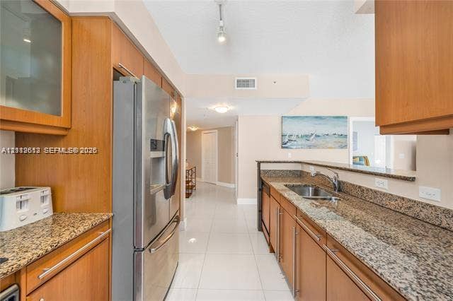 Kitchen featuring wood cabinetry, stone countertops, stainless steel refrigerator, tile flooring, and integrated sink