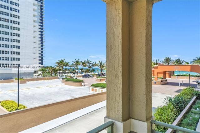 Covered balcony with a textured stucco support pillar, overlooking an exterior surface parking area, landscaped masonry planters, and a coastal landscape