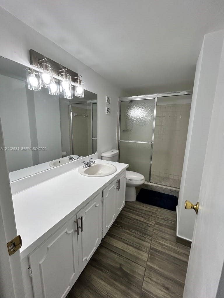 Bathroom featuring a wood-finish tile floor, extended white vanity with a solid-surface countertop, full-width mirror, multi-bulb wall-mounted lighting, and a glass-enclosed shower