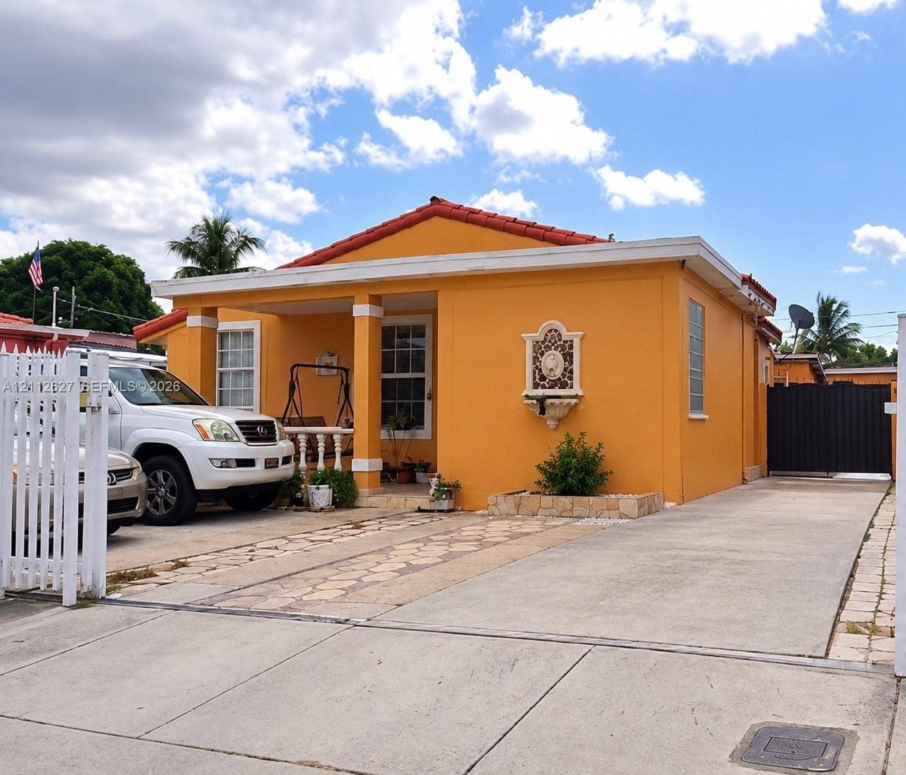 240 E 39th Street, Hialeah, FL 33013 Single-story home featuring a tile roof, covered front porch with square columns, decorative wall-mounted exterior fountain, and a paved driveway with a privacy gate