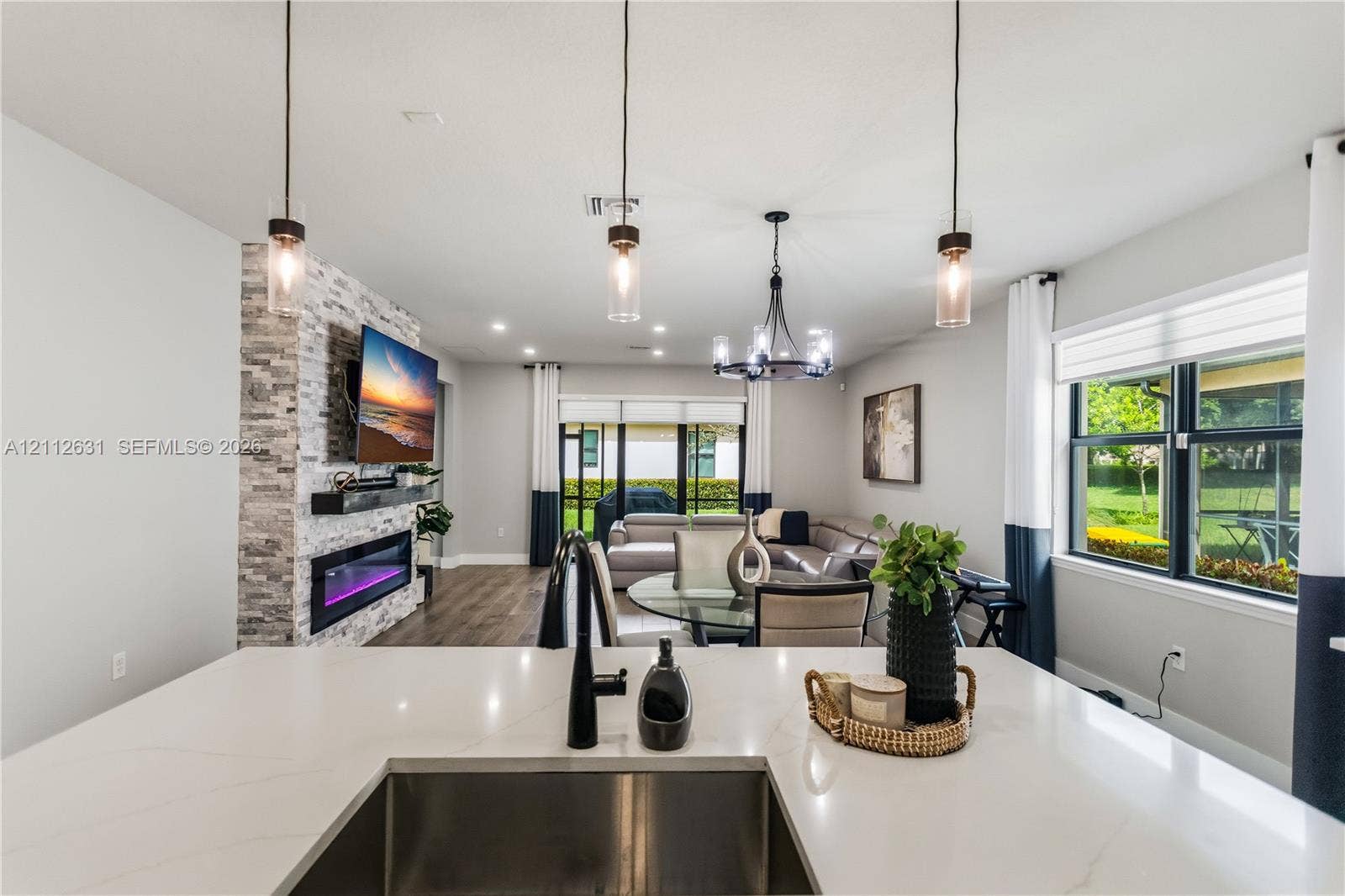 8257 NW 79th Avenue, Tamarac, FL 33321 Kitchen island featuring a white stone countertop, undermount stainless steel sink, and matte black faucet, complemented by a floor-to-ceiling stone-veneer fireplace, wood-finish flooring, and multi-light pendant chandeliers