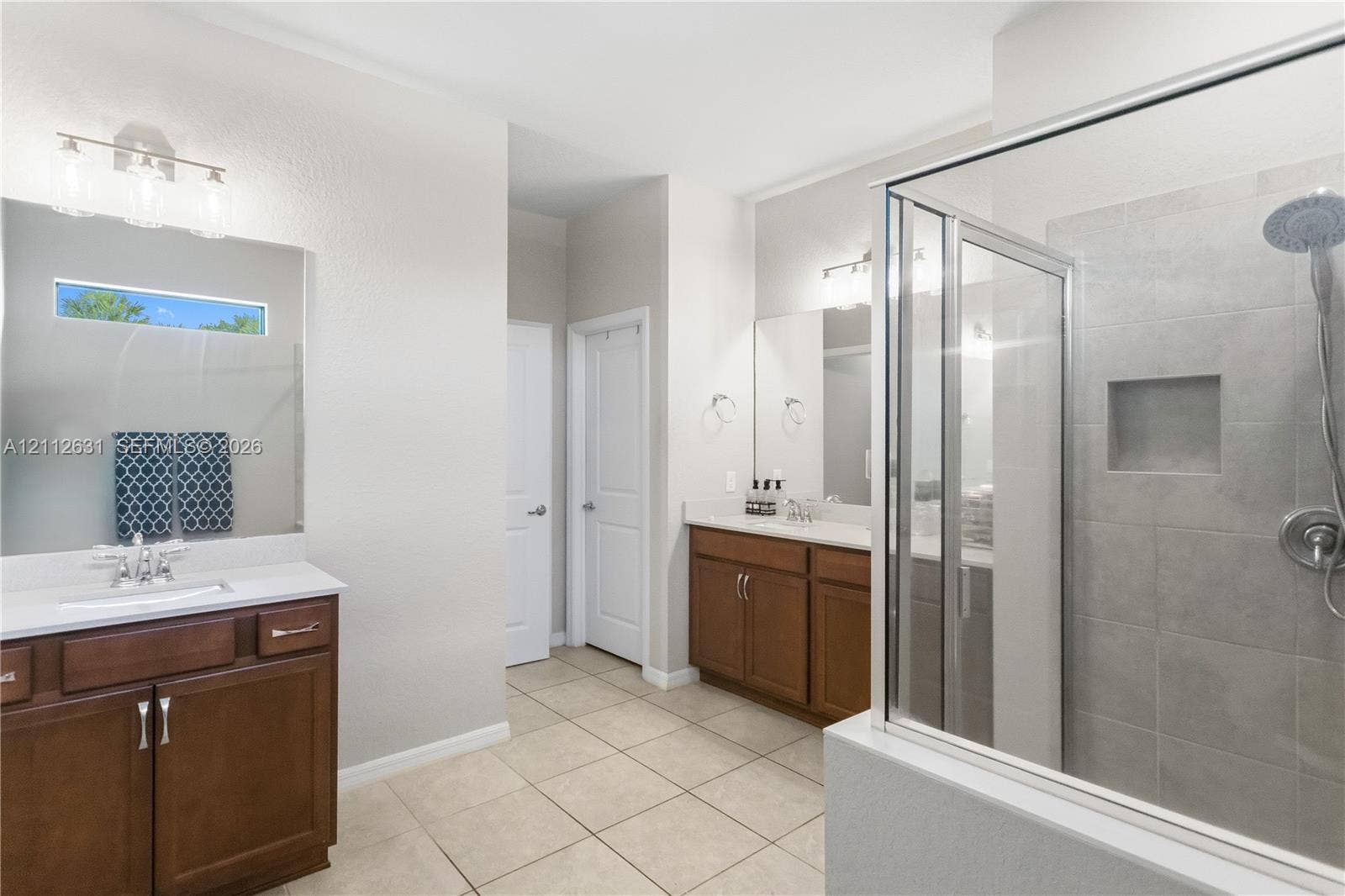8257 NW 79th Avenue, Tamarac, FL 33321 Bathroom featuring dual wood cabinetry vanities with light countertops, large wall-mounted mirrors, tile flooring, and a walk-in shower with tiled surround and built-in recessed niche