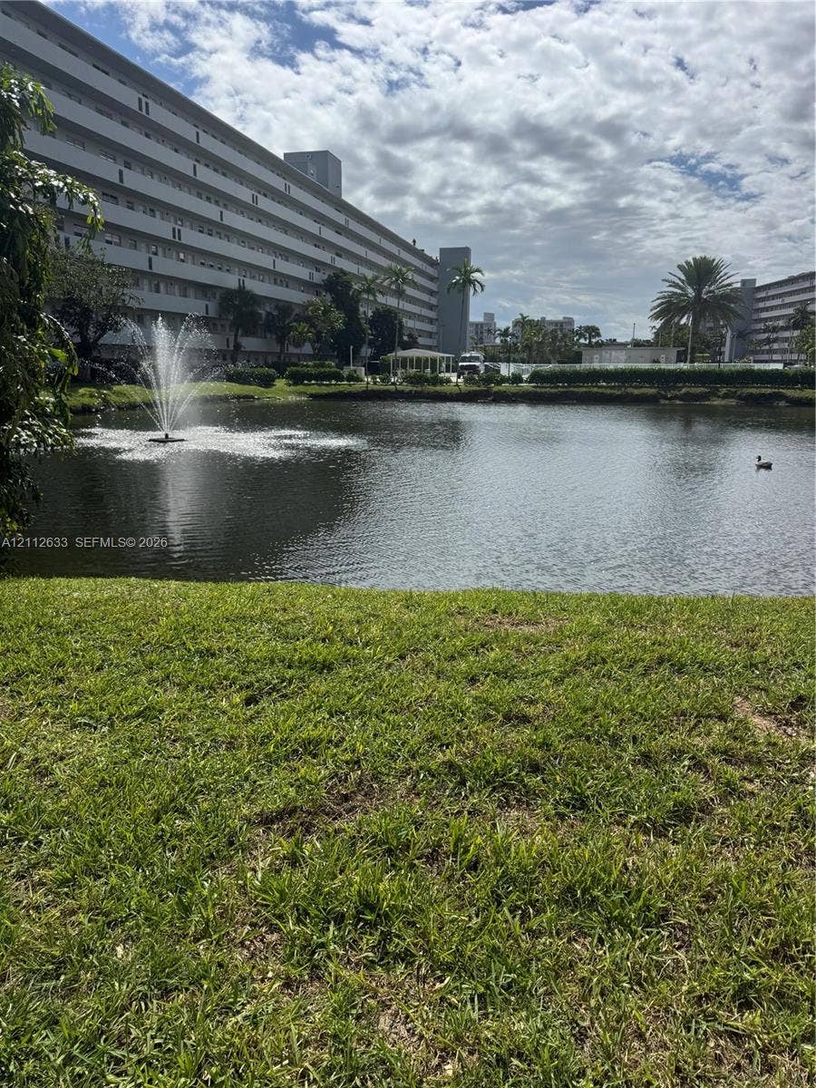 1770 NE 191st Street, #104-1, Miami, FL 33179 Manicured lawn encompassing a central pond with a mechanical fountain, framed by palm trees and multi-story structural exteriors