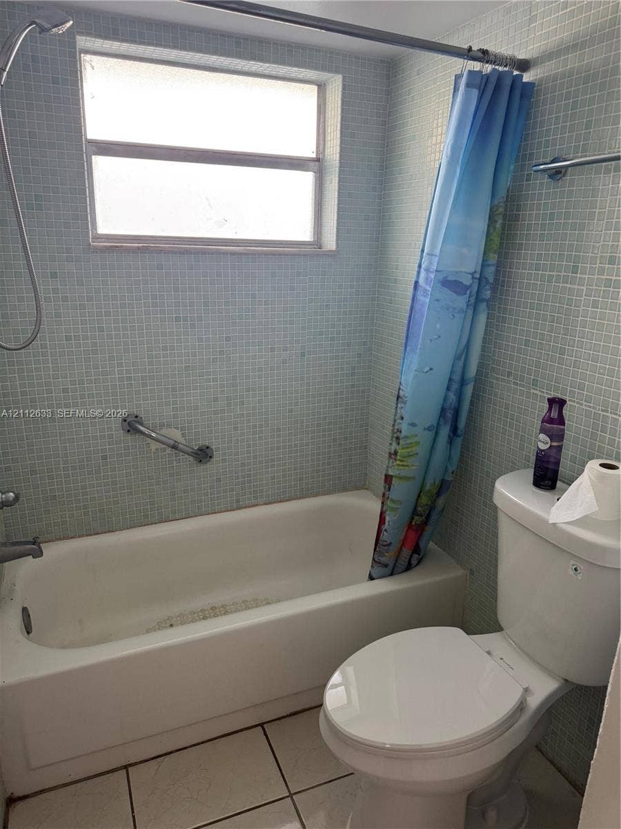 1770 NE 191st Street, #104-1, Miami, FL 33179 Full bathroom featuring light blue mosaic wall tile, white soaking tub with integrated shower head, horizontal window, and white floor tile