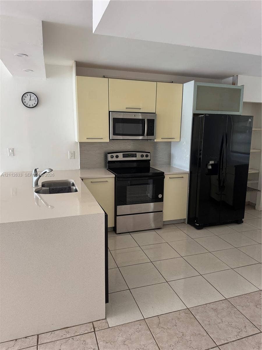 1770 NE 191st Street, #104-1, Miami, FL 33179 Kitchen featuring pale cabinetry, a stainless steel electric range, a microwave, a large-format refrigerator, and a solid-surface peninsula with an integrated sink