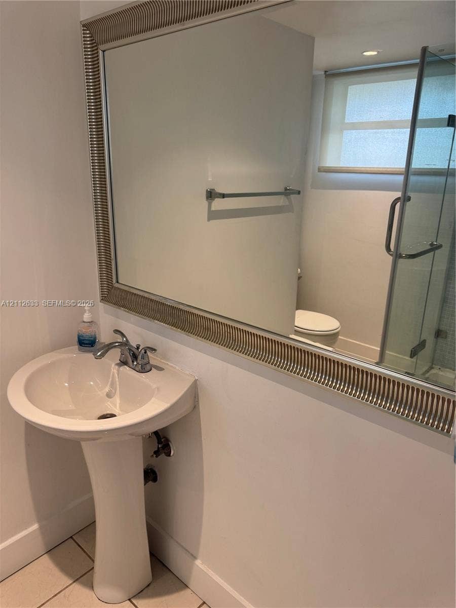 1770 NE 191st Street, #104-1, Miami, FL 33179 Pedestal sink with polished chrome double-handle faucet, large decorative framed mirror, glass-enclosed shower, and light-toned tile flooring