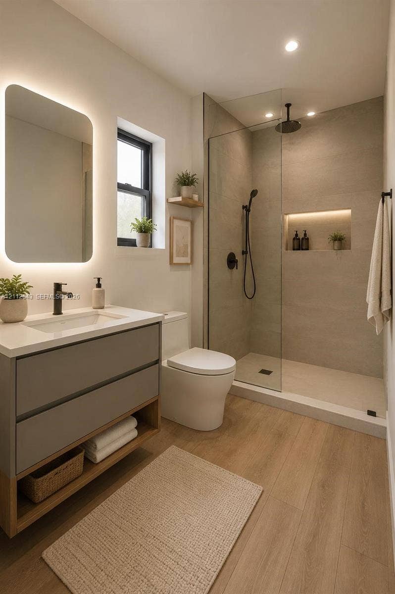 1190 NW 57th Street, Miami, FL 33127 Modern bathroom featuring a gray dual-drawer floating vanity with a white countertop, backlit mirror, wood-finish flooring, and a walk-in shower with a glass enclosure, matte black rainfall showerhead, and integrated recessed shelving