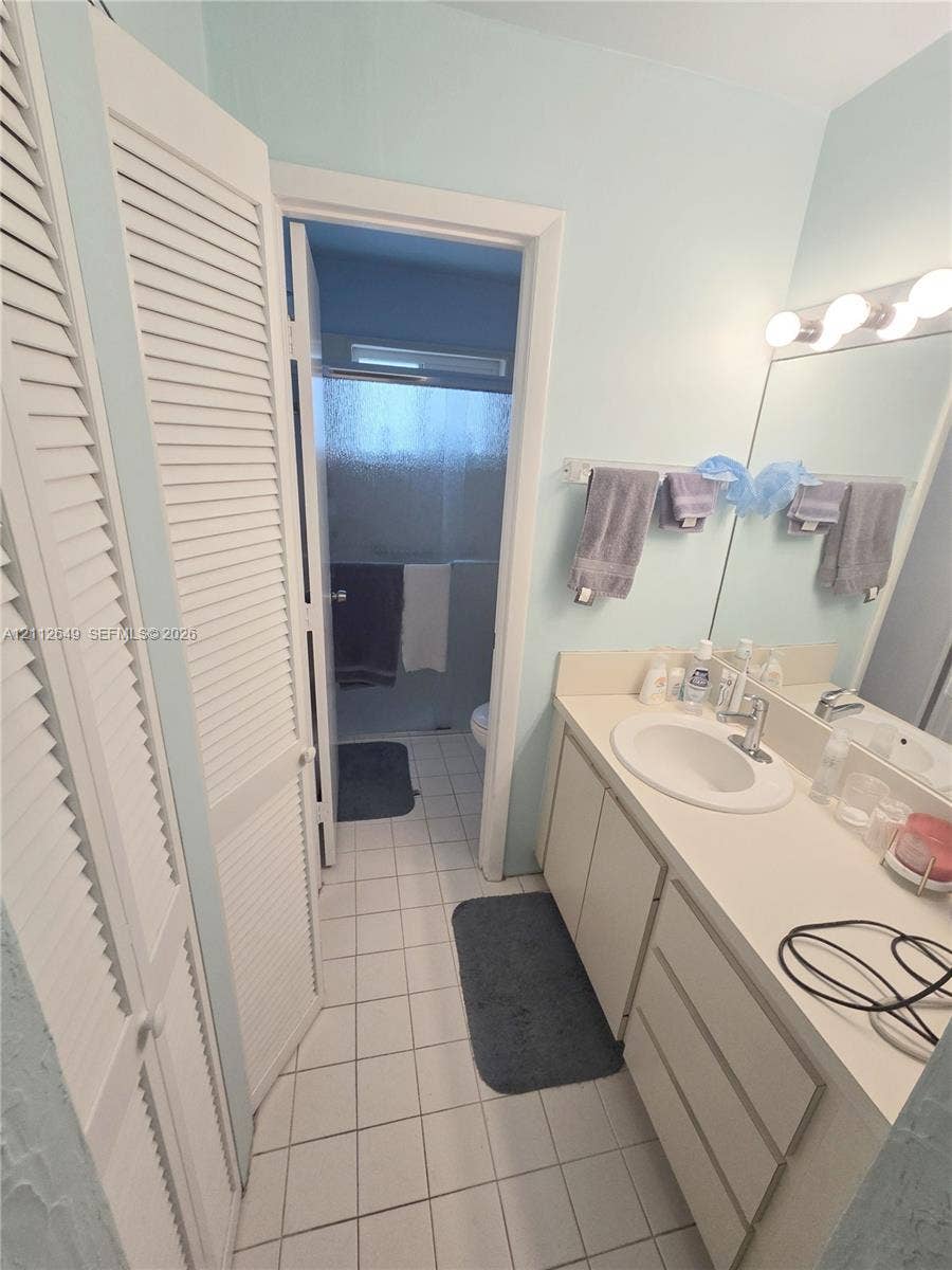 Bathroom featuring a single-sink vanity with white cabinetry, louvered closet doors, tile flooring, and a wall-mounted light fixture above a large mirror