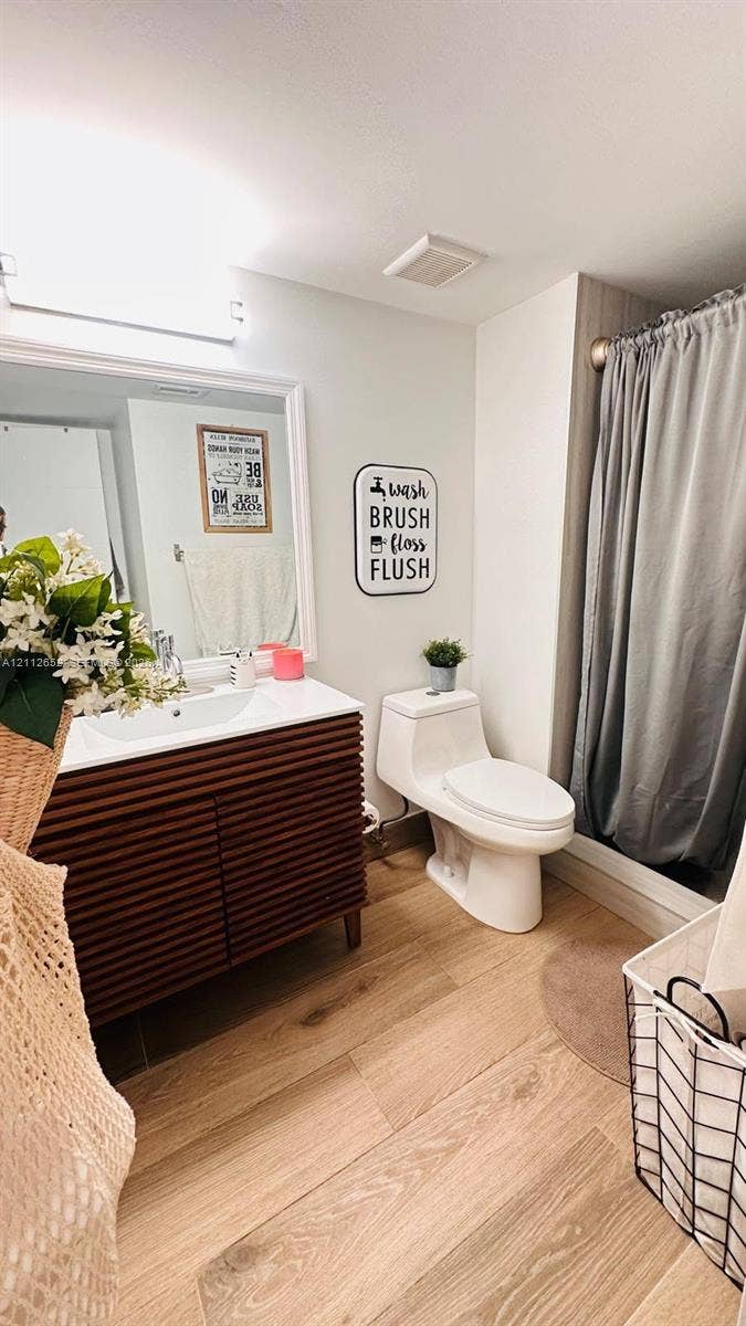 Bathroom featuring a wood-finish vanity with a white countertop and integrated sink, a rectangular framed mirror, a one-piece toilet, and wood-finish flooring
