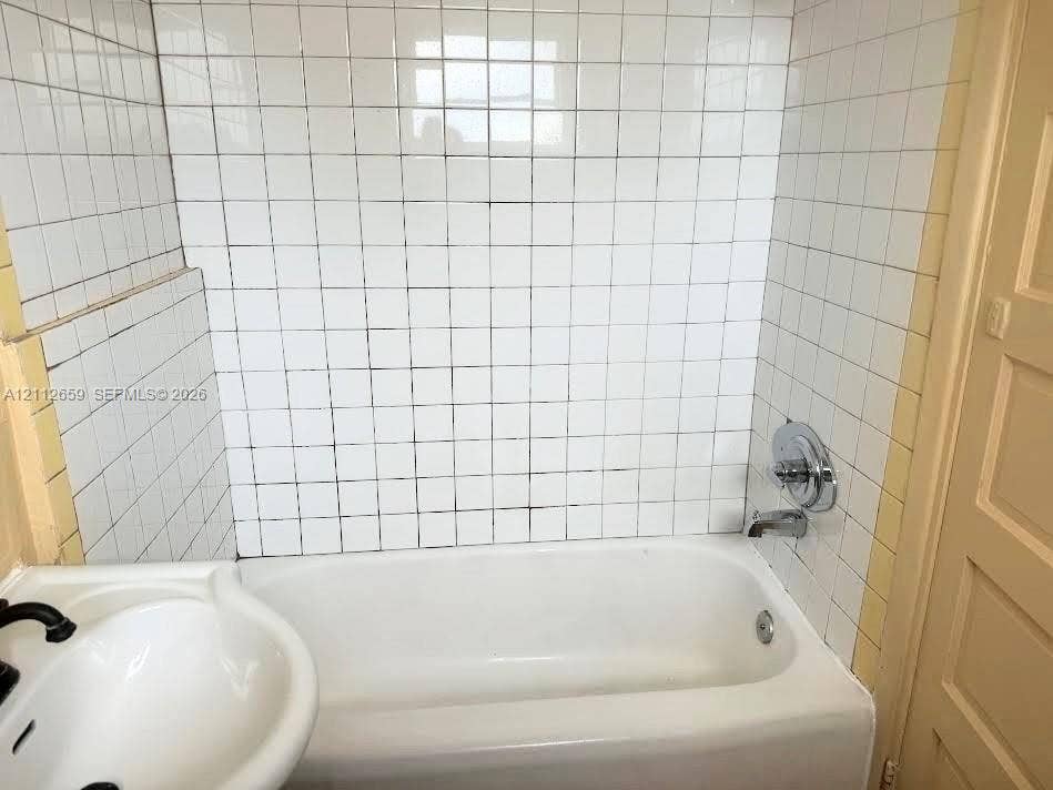 Bathroom featuring a white porcelain tub, square wall tiling, chrome faucet and shower controls, and a pedestal sink with black fixtures
