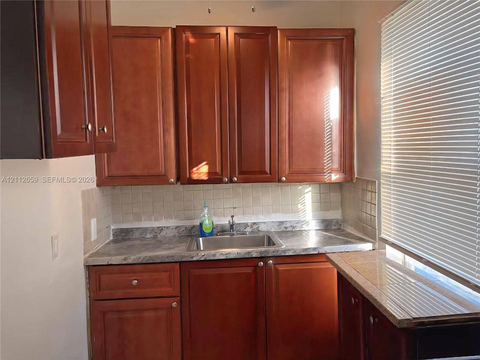 Kitchen area featuring wood-finish cabinetry, stone-patterned laminate countertops, square tile backsplash, and a drop-in stainless steel sink