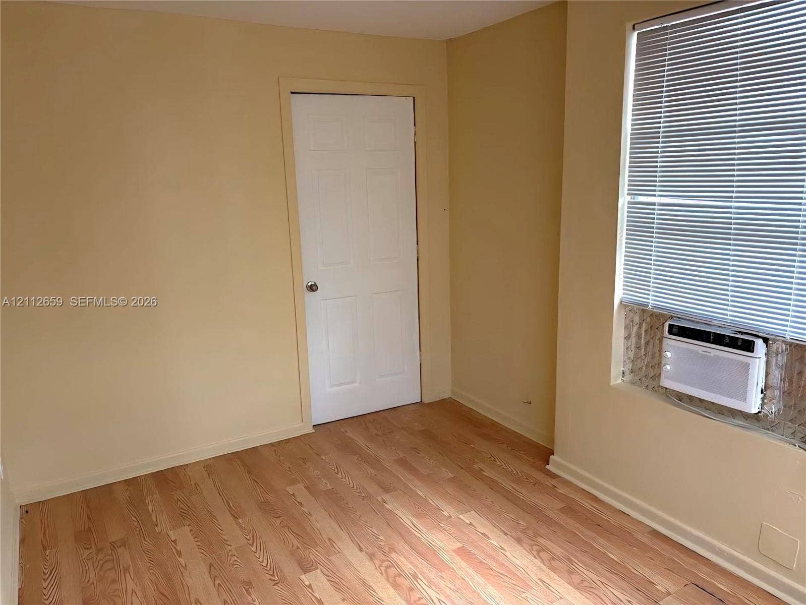 Room featuring wood-finish flooring, a six-panel door, and a wall-mounted air conditioning unit set within a window with horizontal blinds