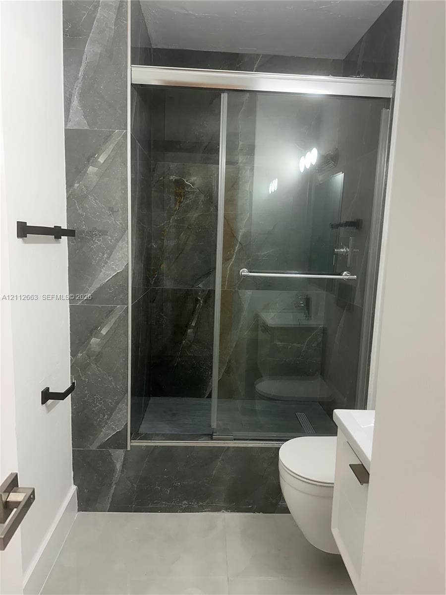 10185 Collins Avenue, #523, Bal Harbour, Bal Harbour, FL 33154 Walk-in shower featuring floor-to-ceiling large-format tile, sliding glass door, wall-mounted matte black hardware, and an elongated toilet