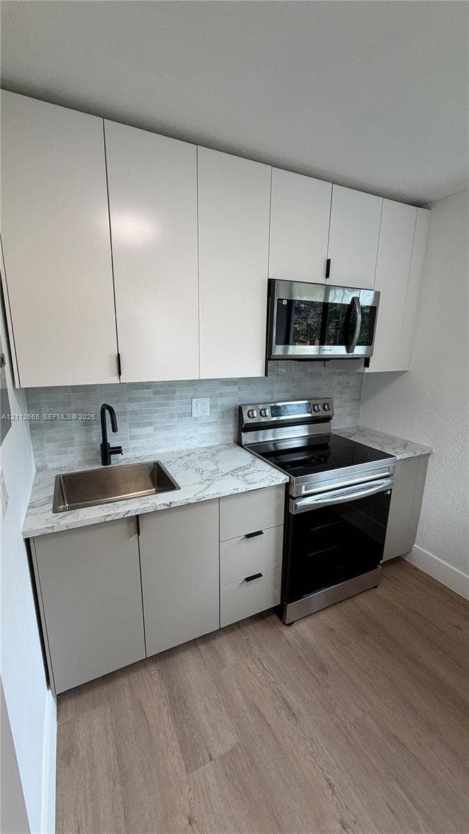 3314 NW 8th, #2, Miami, Miami, FL 33131 Modern kitchen featuring white flat-panel upper cabinetry, light gray base cabinets, patterned stone-finish countertops, stainless steel appliances, and wood-finish flooring