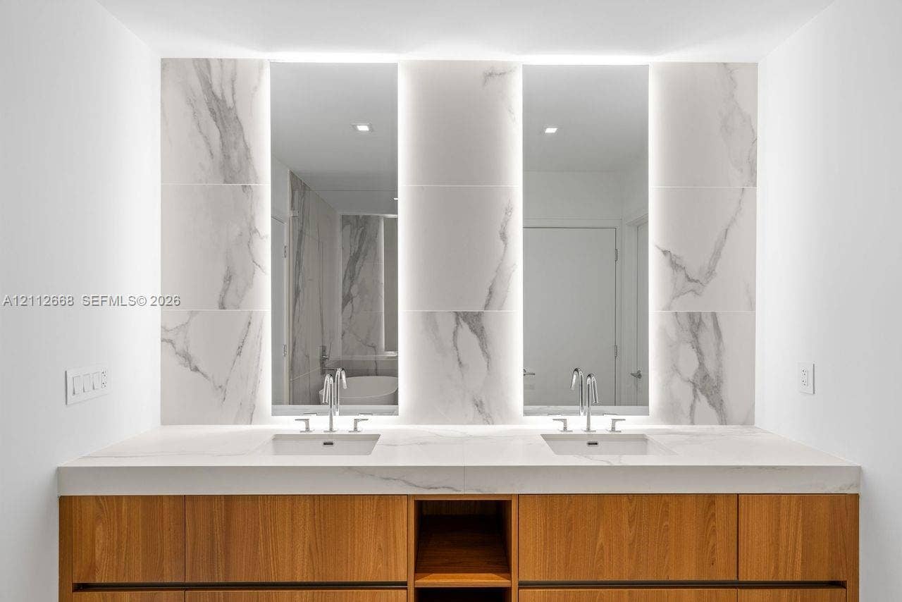1400 Biscayne Boulevard, #1803, Miami, Miami, FL 33132 Double vanity featuring a wood-finish cabinet, stone-surfaced countertop, integrated rectangular sinks, backlit vertical mirrors, and stone-tiled backsplash