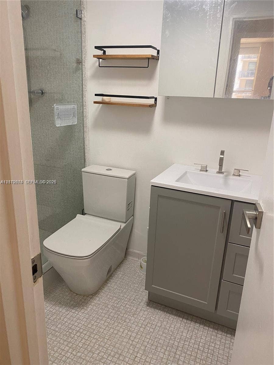 Bathroom featuring a gray vanity cabinet with an integrated white sink, wall-mounted mirrored medicine cabinet, glass-enclosed shower, dual tiered wood and metal wall shelving, and tile flooring