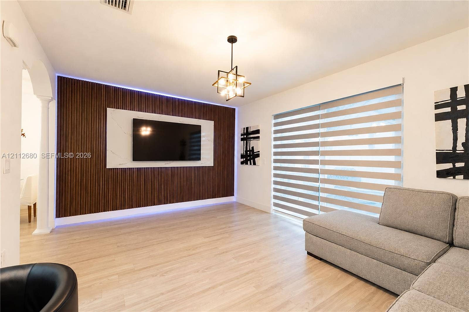 890 SW 154th Path, Miami, FL 33194 Vertical wood-slat accent wall with integrated LED lighting, stone-finish media panel, wood-finish flooring, geometric multi-bulb chandelier, and large dual-pane horizontal blind window covering