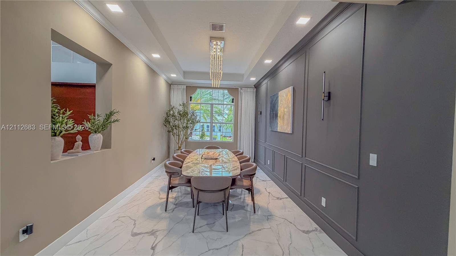 Dining area featuring large-format tile flooring, a tray ceiling with a linear crystal chandelier, an accent wall with wainscoting, and a recessed architectural wall niche