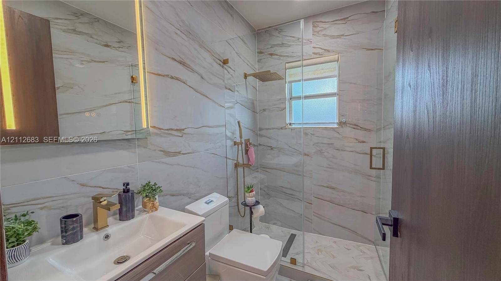 Bathroom featuring large-format wall and floor tile, glass-enclosed shower with gold-tone fixtures, wall-mounted vanity, and integrated vertical lighting