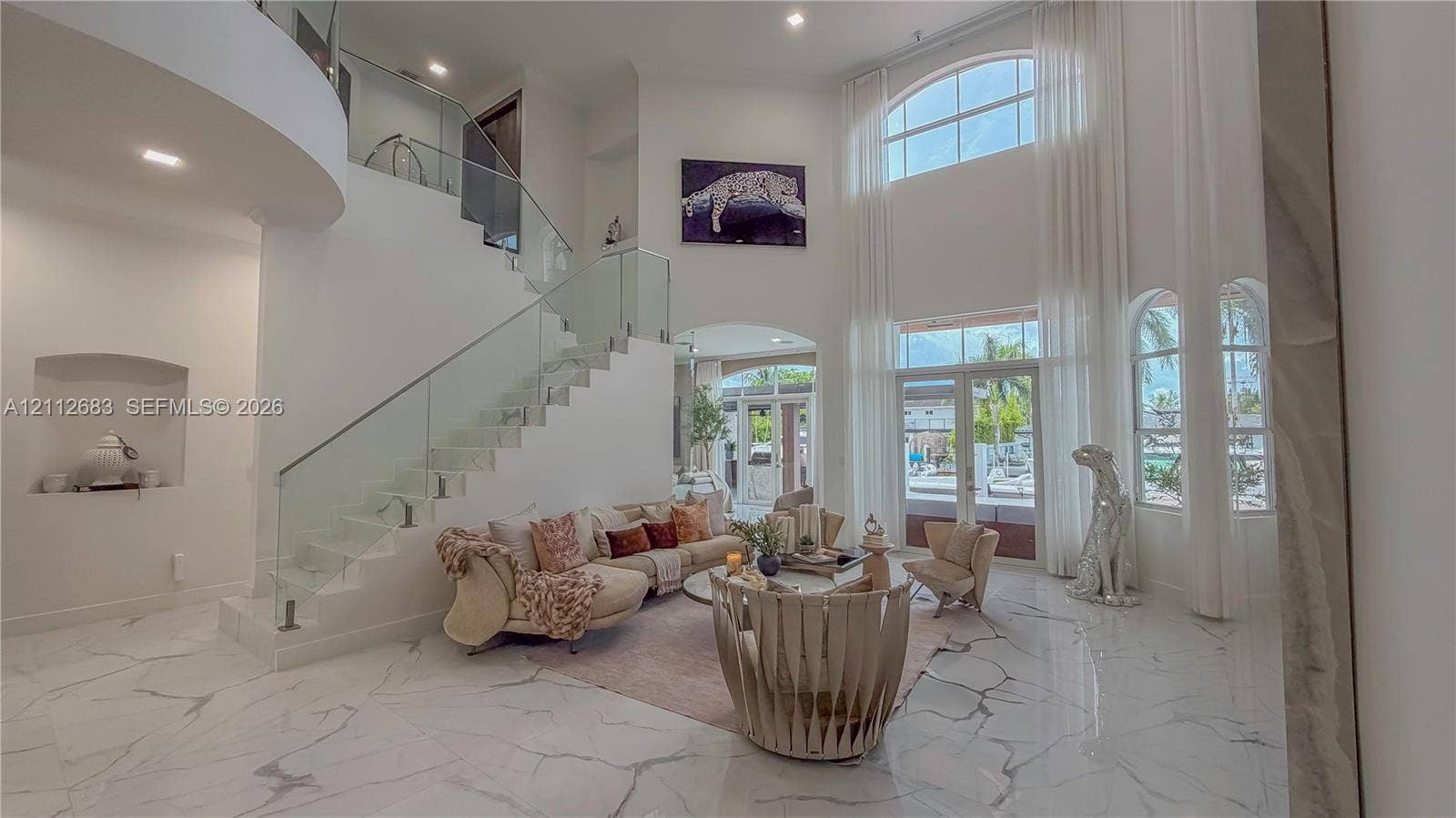 Grand foyer featuring high ceilings, large-format stone flooring, a sweeping staircase with a glass panel railing, arched windows, and a built-in recessed wall niche