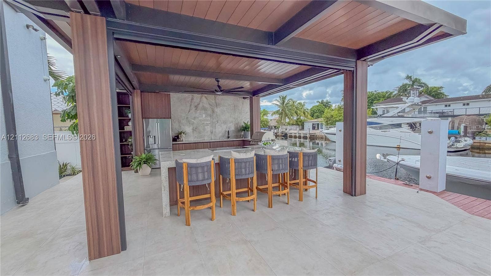 Covered outdoor kitchen featuring a stone-topped island with integrated seating, natural wood-finish ceiling accents, built-in stainless steel appliances, and large-format tile flooring