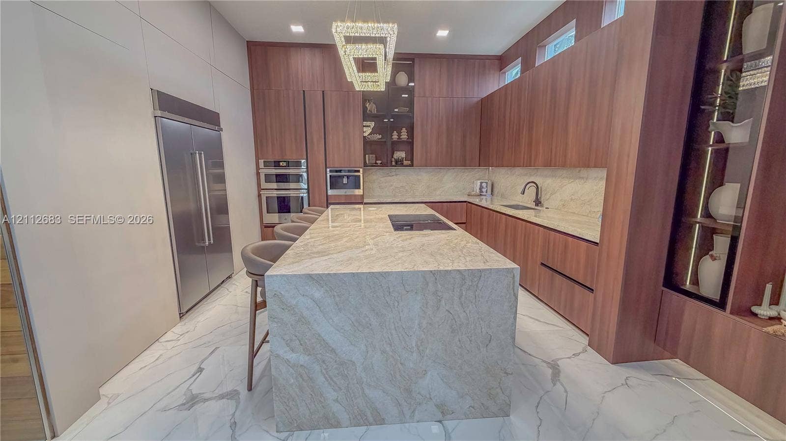 Kitchen featuring wood-finish cabinetry, a waterfall-edge stone island with an integrated cooktop, stone backsplash, built-in stainless steel appliances, and large-format tile flooring