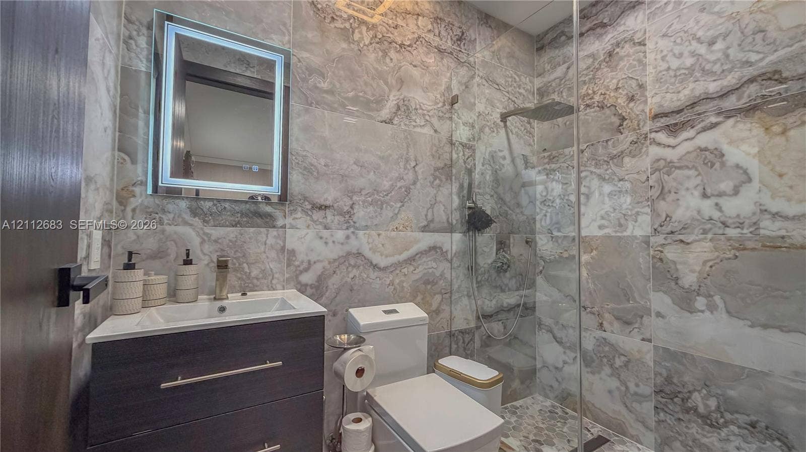 Bathroom featuring floor-to-ceiling large-format natural stone wall tile, vanity with dark wood-finish cabinetry, frameless glass walk-in shower enclosure, rainfall showerhead, and integrated LED-backlit mirror