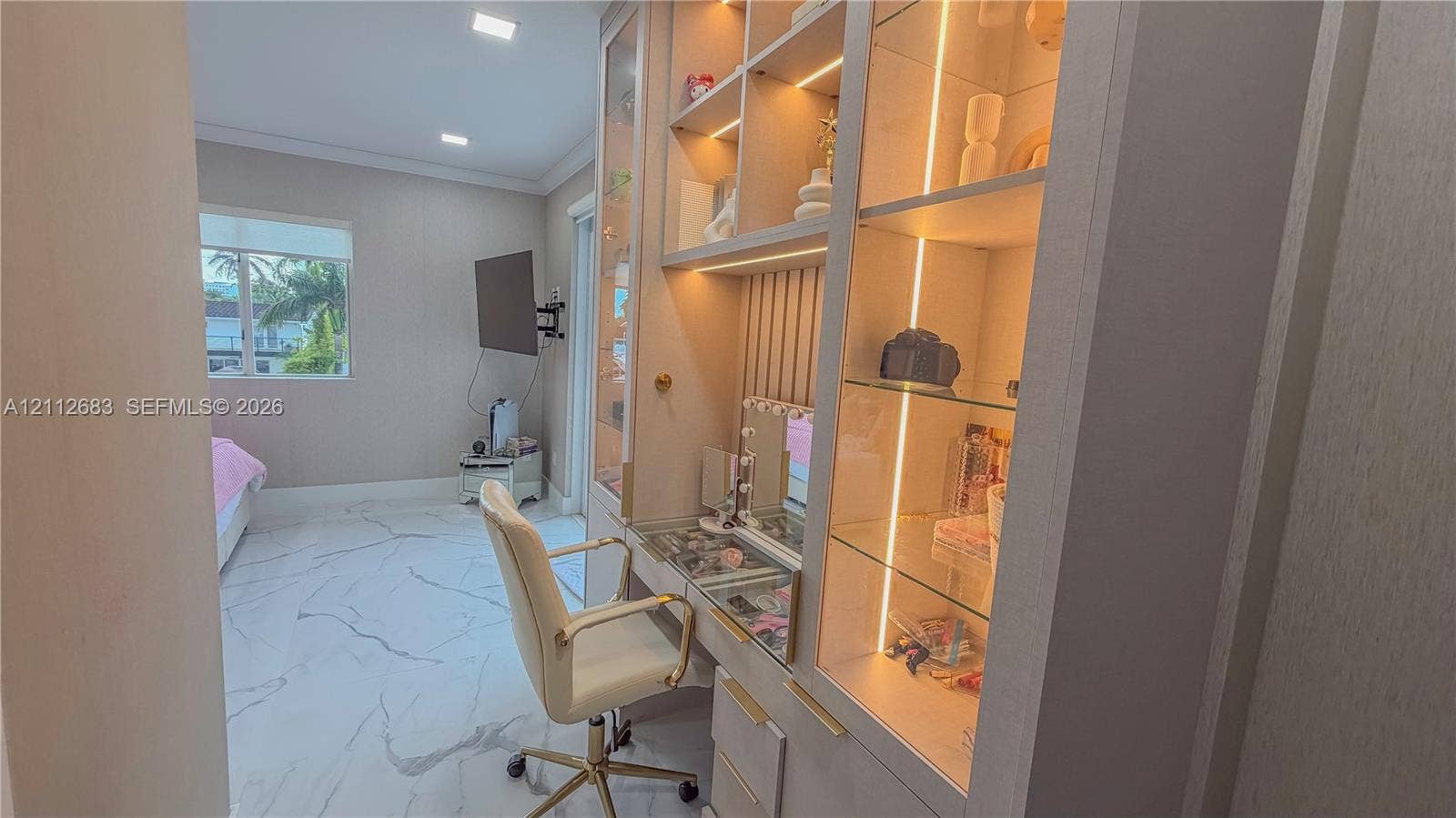 Custom floor-to-ceiling built-in cabinetry featuring integrated glass display shelving, recessed LED lighting, and a dedicated vanity workspace with light-toned cabinetry, set against stone-finish tile flooring