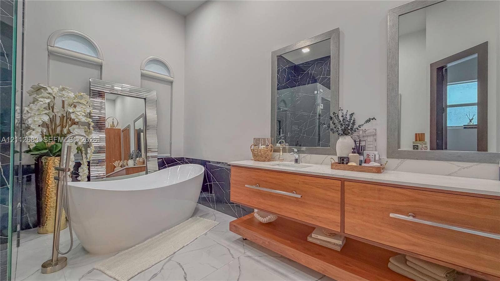 Freestanding soaking tub with floor-mounted faucet, dual-sink floating vanity with wood cabinetry, quartz countertop, and large wall-mounted mirrors