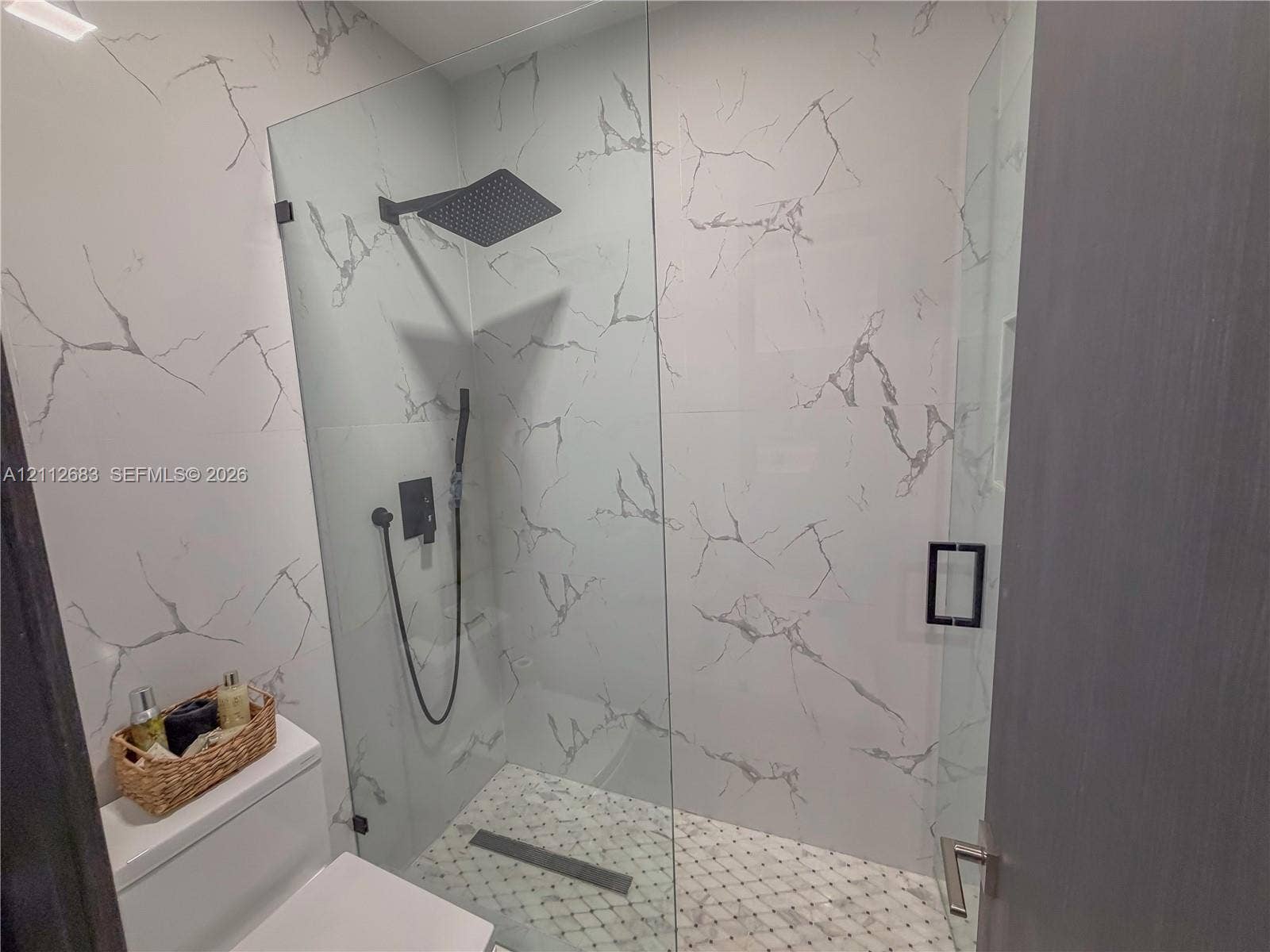 Walk-in shower featuring floor-to-ceiling stone-finish tile, frameless glass enclosure, matte black rain-style shower head with handheld attachment, and recessed wall niche