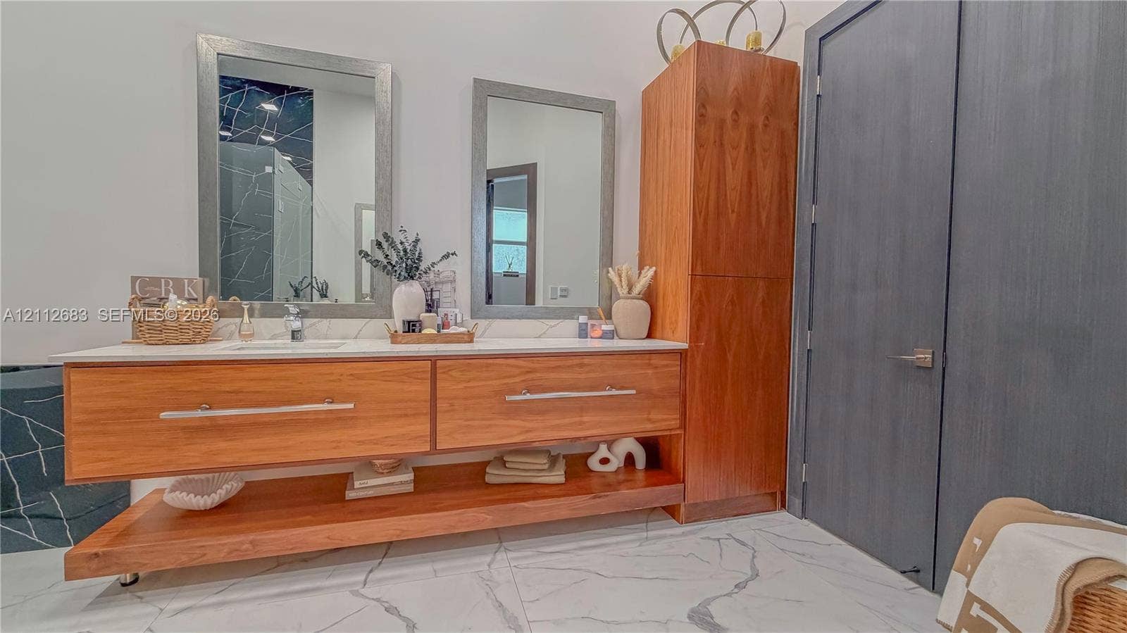 Floating vanity featuring natural wood-finish cabinetry, a white countertop with dual under-mount sinks, integrated vertical storage, and dual framed wall mirrors over large-format veined tile flooring