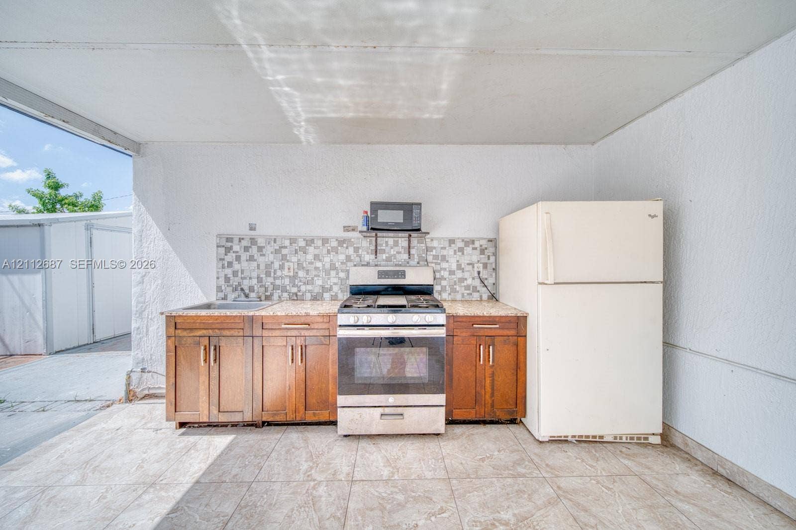 9340 SW 26th Street, Miami, FL 33165 Outdoor kitchen featuring wood-finish cabinetry, stone-look countertop, tiled backsplash, stainless steel gas range, and refrigerator