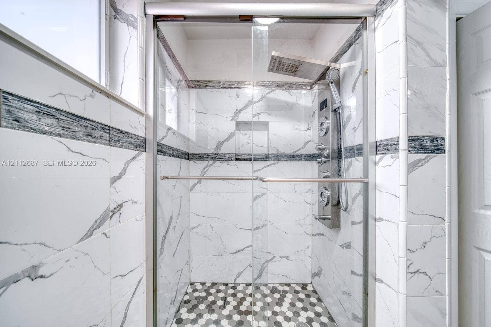 9340 SW 26th Street, Miami, FL 33165 Walk-in shower with floor-to-ceiling veined tile, integrated recessed shelving, multi-head rainfall shower panel, and hexagonal mosaic tile flooring