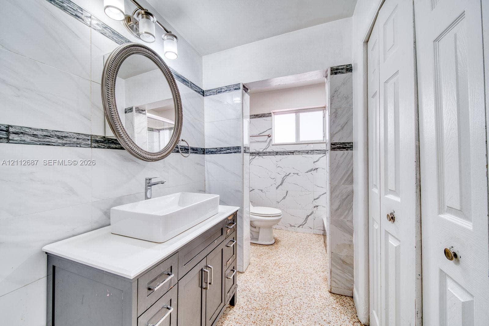 9340 SW 26th Street, Miami, FL 33165 Bathroom featuring a floating vanity with a rectangular vessel sink, polished chrome faucet, wall-mounted oval mirror, and full-height stone-finish wall tile with decorative accent inlay