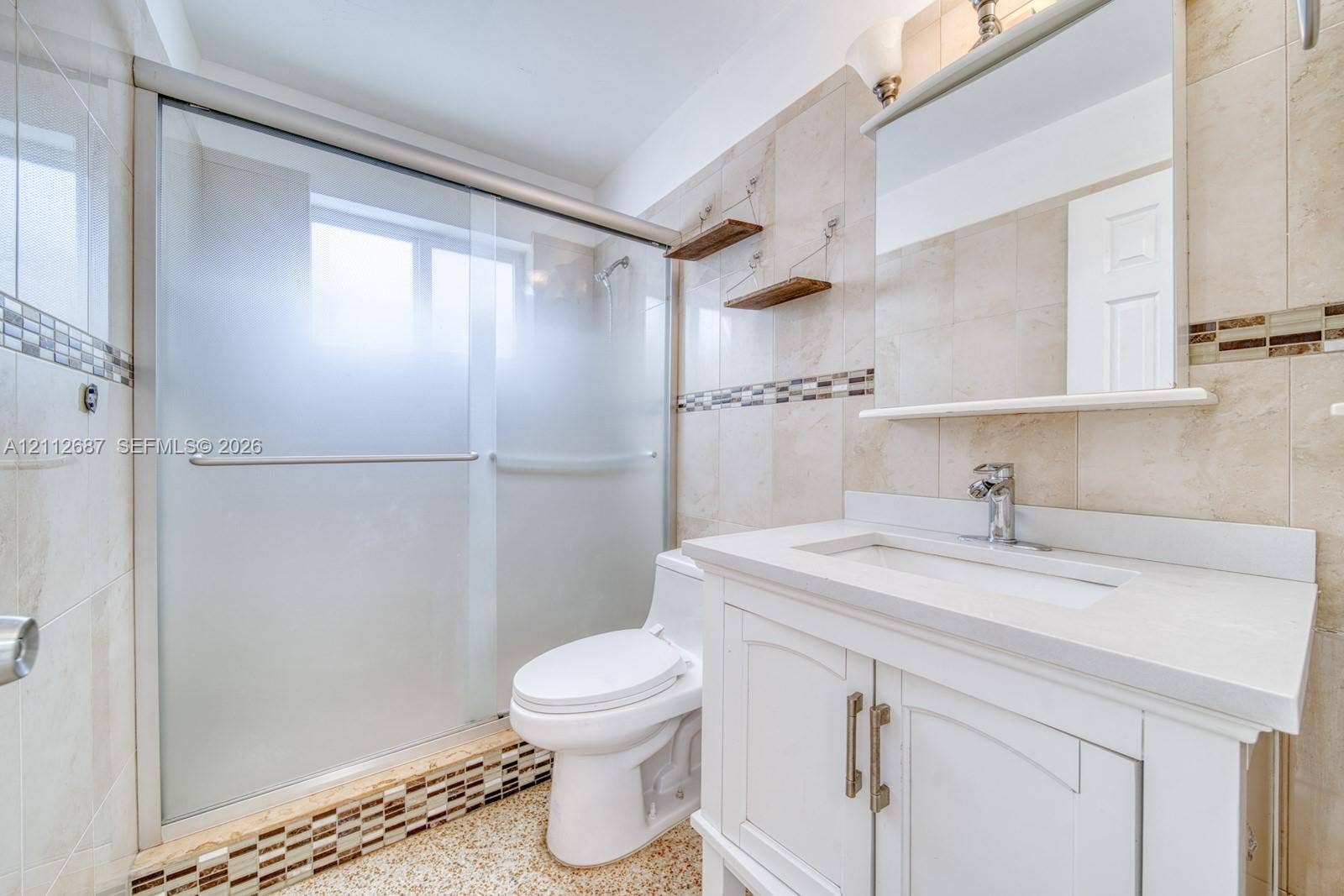 9340 SW 26th Street, Miami, FL 33165 Bathroom featuring a white vanity with a composite countertop, tiled shower enclosure with sliding frosted glass doors, stone-tiled walls with mosaic inlay, and wall-mounted shelving