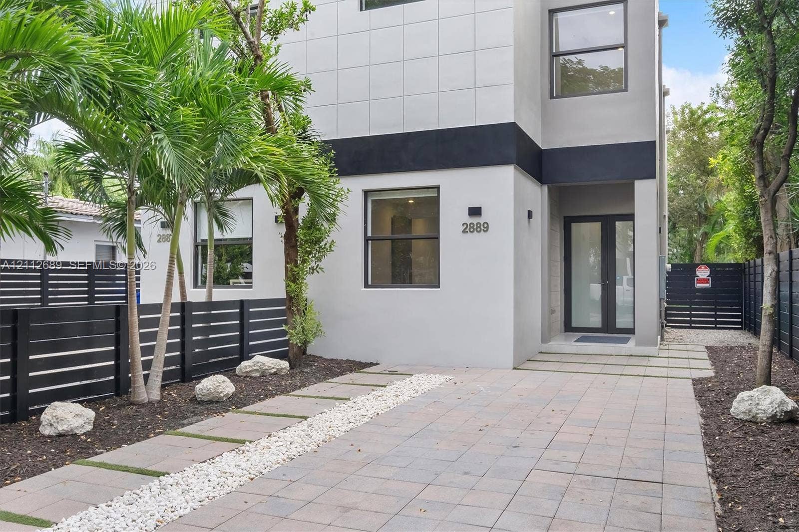 2889 SW 33rd Avenue, #2889, Miami, Miami, FL 33133 Modern two-story exterior featuring smooth stucco siding, large rectangular windows, black horizontal perimeter fencing, paver driveway, and a glass-paneled entry door