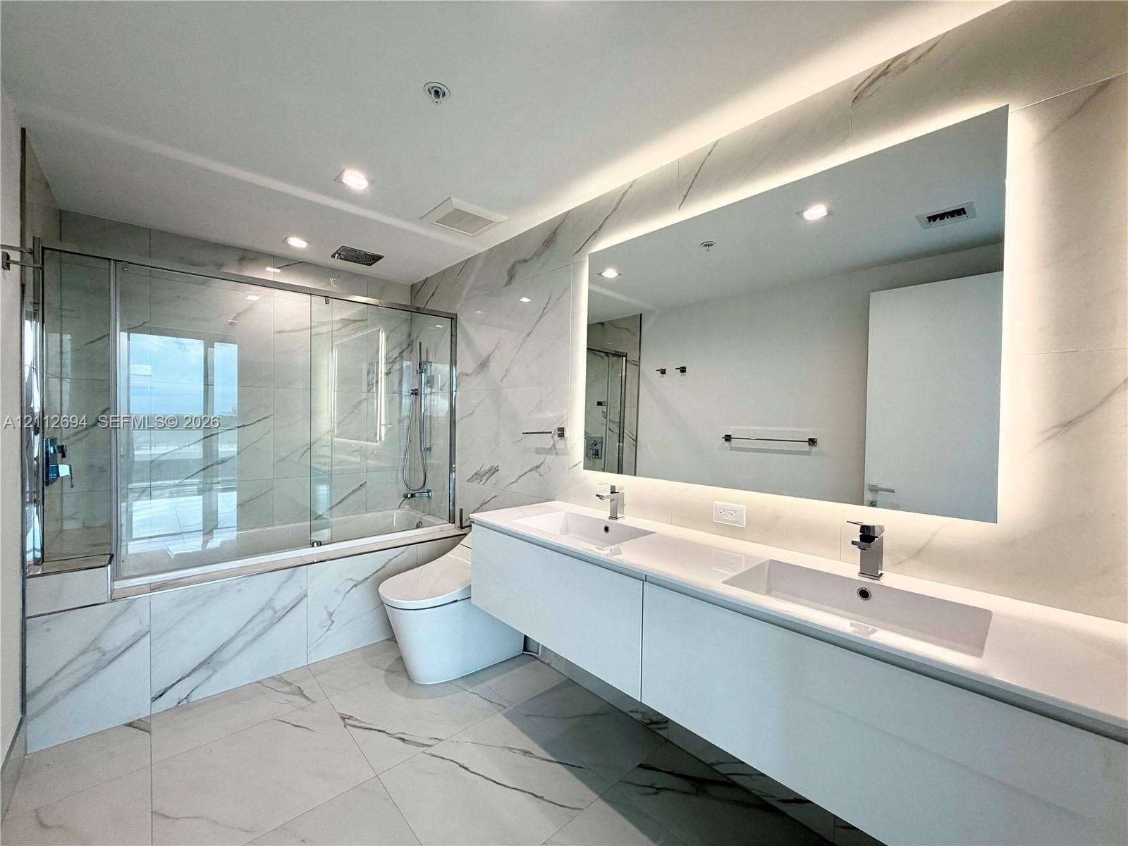 1170 E Hallandale Beach Boulevard, #503, Hallandale Beach, Hallandale Beach, FL 33009 Double floating vanity with integrated rectangular sinks, expansive LED-backlit mirror, large-format stone-finish floor and wall tiles, and glass-enclosed bathtub with chrome fixtures