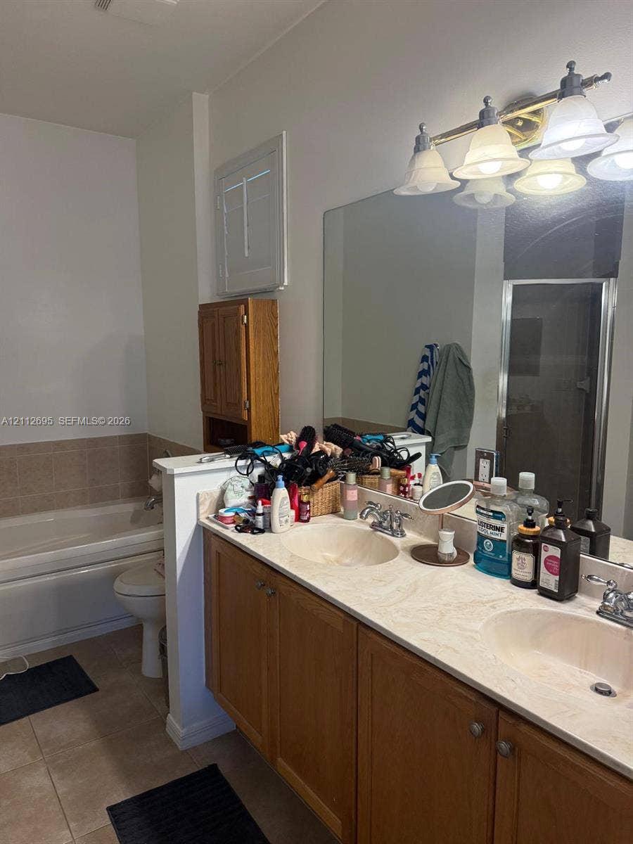 9812 SW Glenbrook Drive, Port St Lucie, FL 34987 Dual-sink vanity with a solid-surface countertop and light-wood cabinetry, large wall-mounted mirror, four-light vanity fixture, built-in medicine cabinet, and a tiled soaking tub