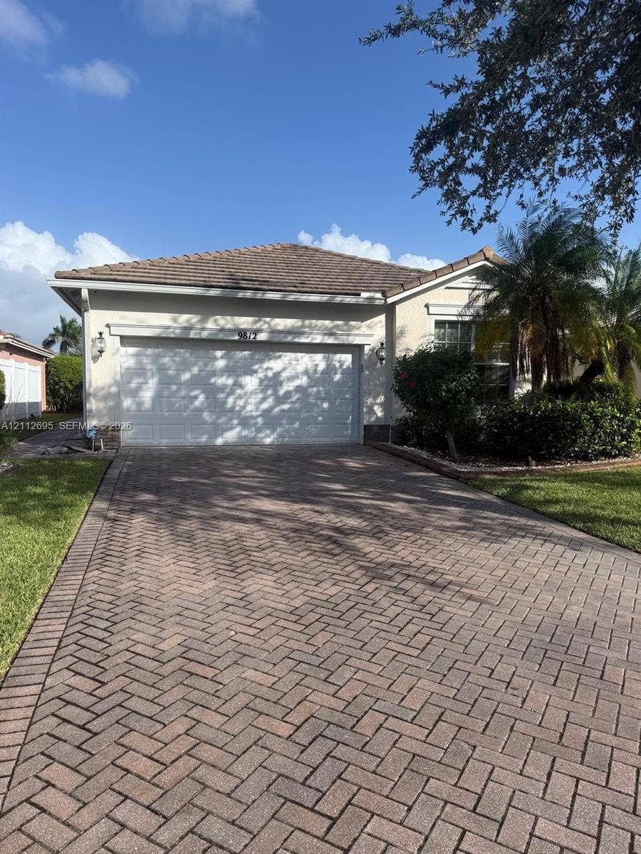 9812 SW Glenbrook Drive, Port St Lucie, FL 34987 Stucco exterior featuring a tiled roof, herringbone-patterned paver driveway, two-car garage, and wall-mounted coach lighting