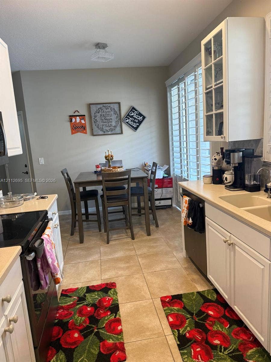9812 SW Glenbrook Drive, Port St Lucie, FL 34987 Kitchen featuring white shaker-style cabinetry, a glass-front upper cabinet, solid surface countertops, dual-basin sink, and ceramic tile flooring