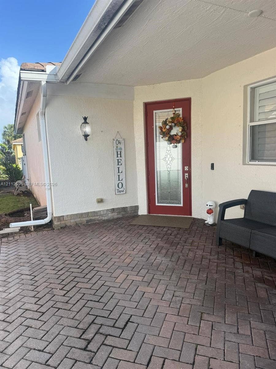 9812 SW Glenbrook Drive, Port St Lucie, FL 34987 Covered entryway featuring brick-paver flooring in a herringbone pattern, exterior wall-mounted light fixture, and a decorative entry door with an integrated glass panel