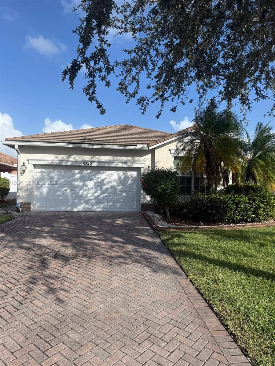 9812 SW Glenbrook Drive, Port St Lucie, FL 34987 Stucco exterior featuring a tiled hip roof, two-car garage, herringbone-patterned paver driveway, and manicured landscaping