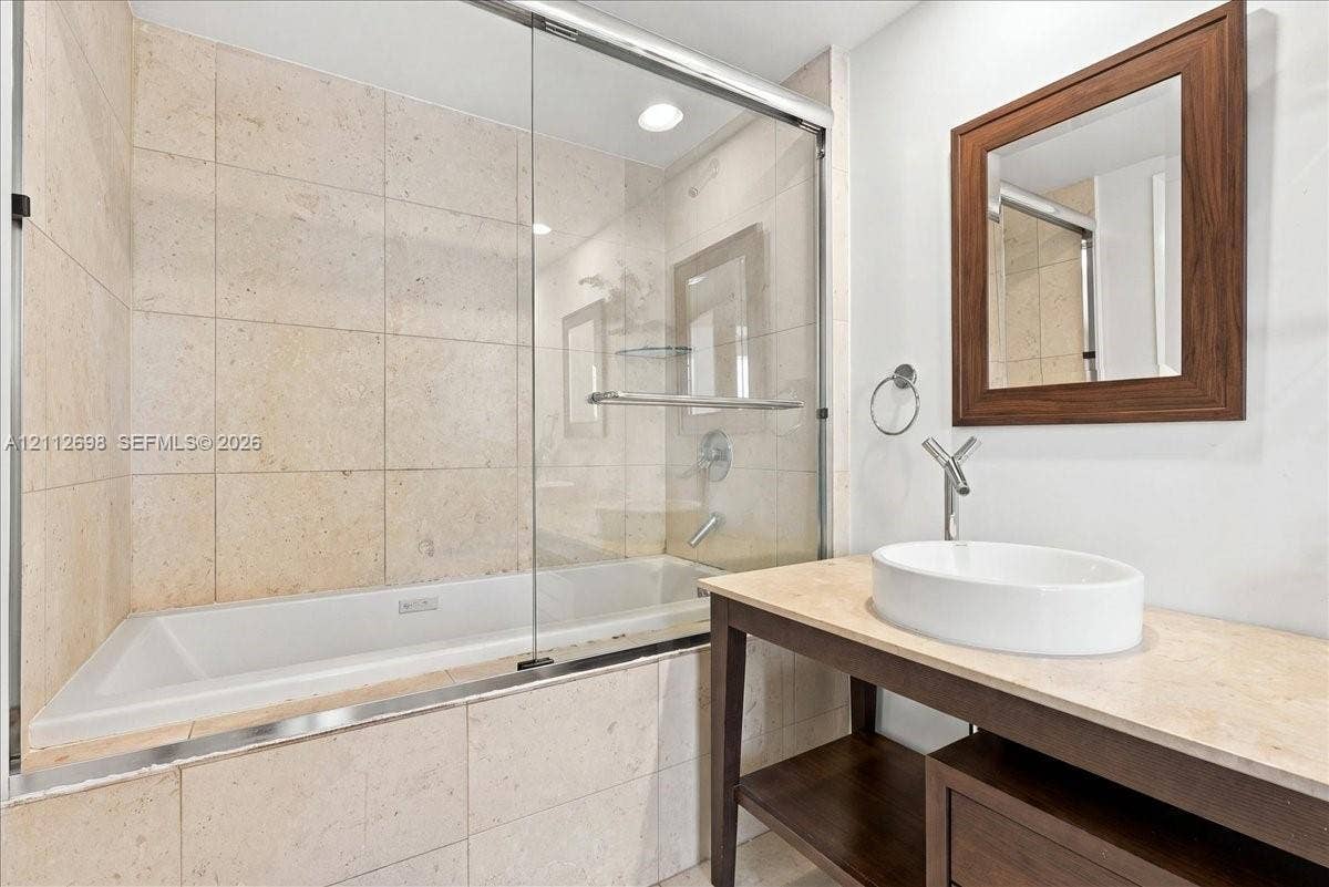 475 Brickell Avenue, #4511, Miami, FL 33131 Bathroom featuring a stone-tiled bathtub enclosure with a glass shower door, open-base vanity with a stone countertop, oval vessel sink, and a wood-framed wall mirror