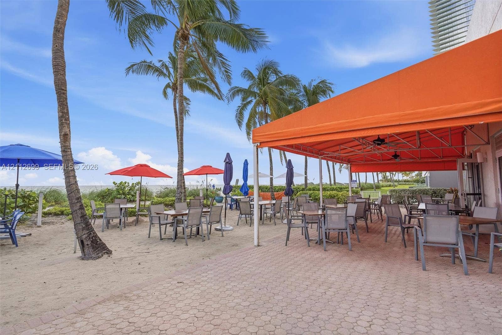 19111 Collins Avenue, #505, Sunny Isles Beach, FL 33160 Expansive paver patio featuring a large fixed orange awning, direct sandy beach access, mature palm trees, and ceiling-mounted fans