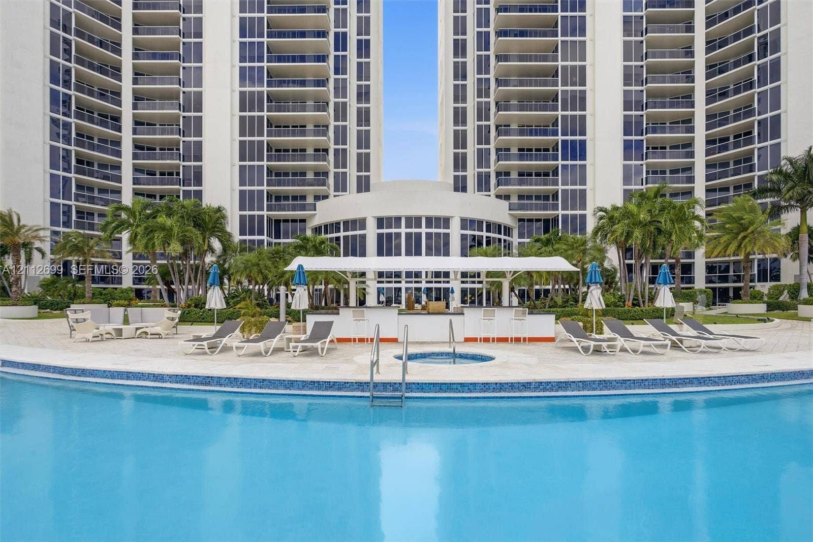 19111 Collins Avenue, #505, Sunny Isles Beach, FL 33160 Curved swimming pool with tile trim, integrated spa, poolside bar under a covered canopy, and paved deck featuring mature palm trees