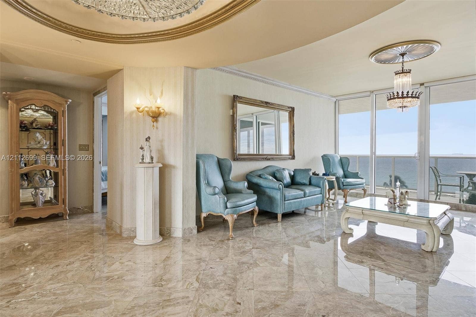 19111 Collins Avenue, #505, Sunny Isles Beach, FL 33160 Polished stone flooring, recessed tray ceiling with gold-tone molding, crystal chandelier, and floor-to-ceiling glass sliding doors with ocean backdrop