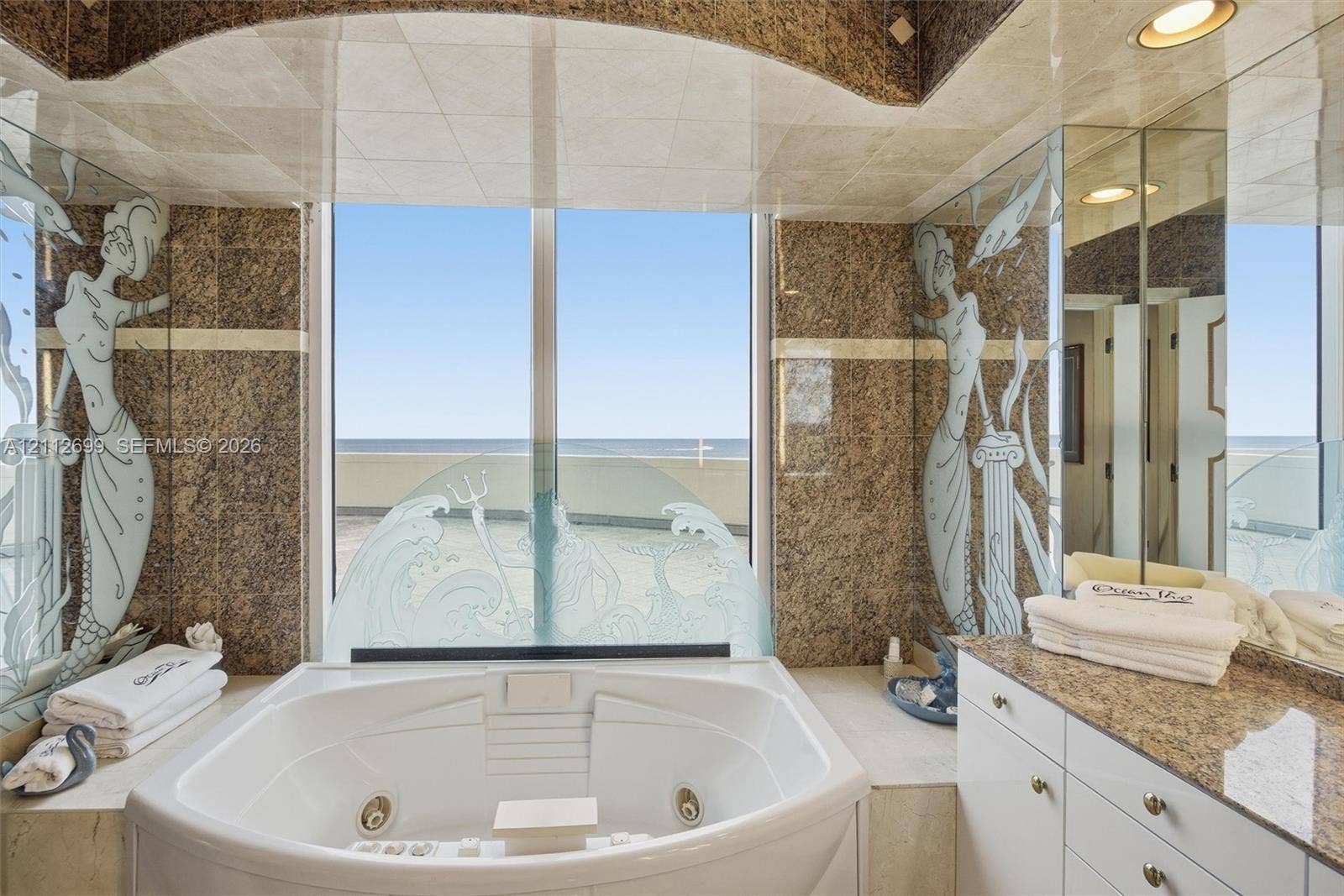 19111 Collins Avenue, #505, Sunny Isles Beach, FL 33160 Custom soaking tub with integrated jets, large window with ocean horizon backdrop, stone wall cladding, decorative etched glass panels, and built-in vanity with stone countertop