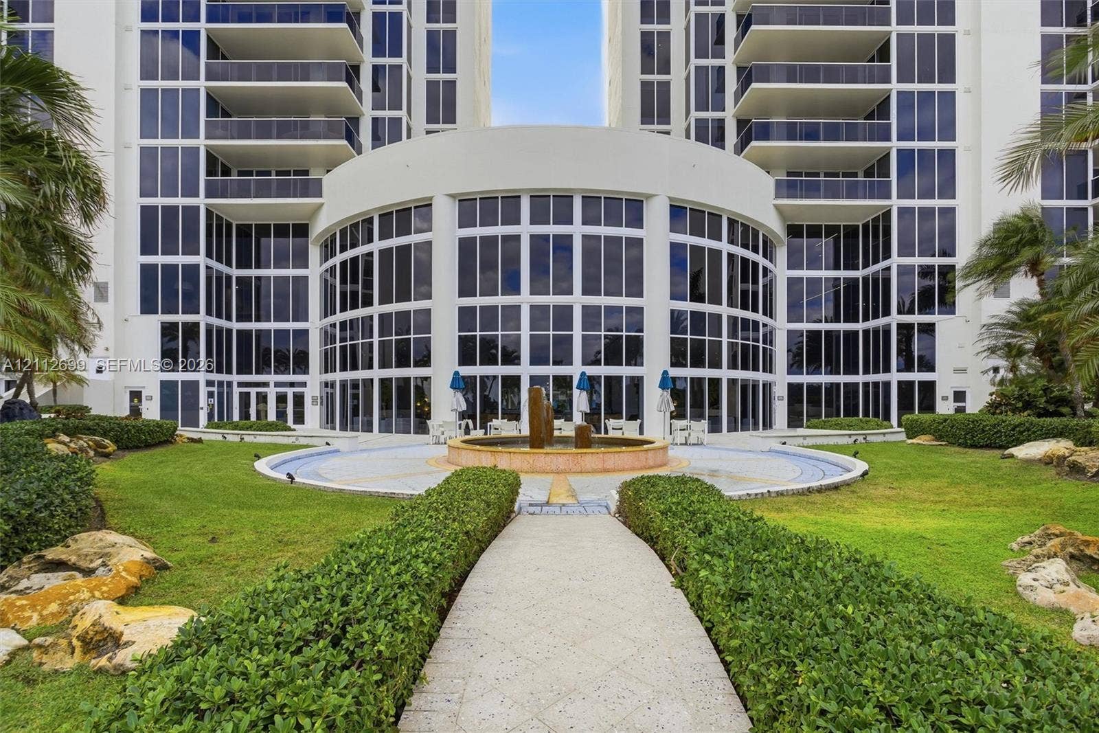 19111 Collins Avenue, #505, Sunny Isles Beach, FL 33160 High-rise exterior featuring a multi-story glass facade, private balconies, a central water fountain, stone-paved pathways, and manicured landscaping